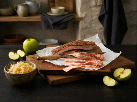 Sliced Opa's Smoked Peppered Bacon on parchment with a knife, green apples, and a bowl of sauerkraut on a dark wooden board.