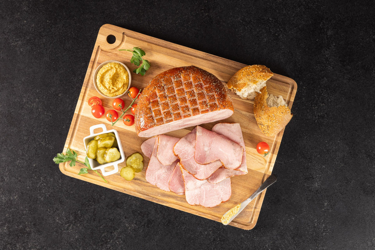 Opa's Boneless Peppered Ham, half sliced, on a cutting board with pickles, mustard and bread